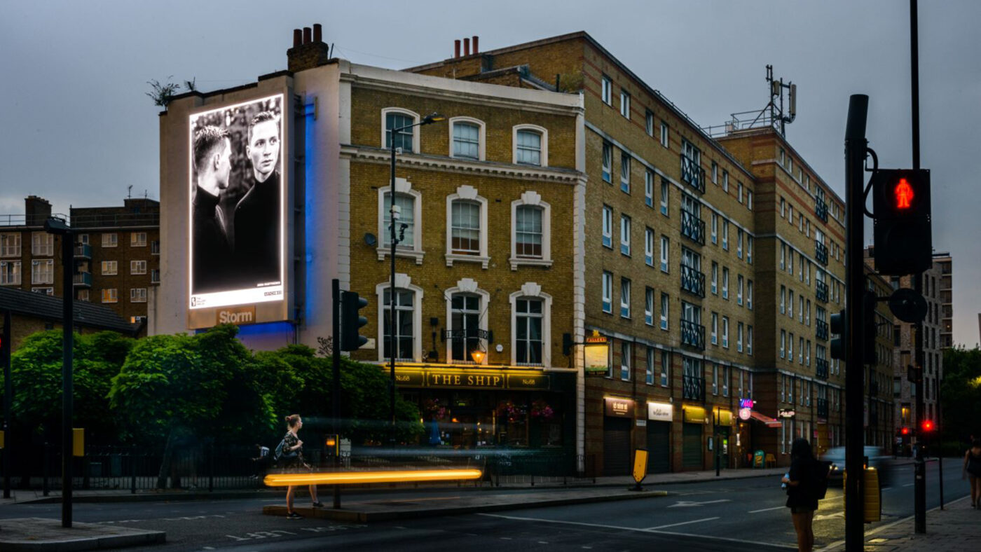 A photograph of a London street with a billboard on the side of a pub featuring one of the artworks. The artwork is a photograph of two mid-twenties men standing close to each other.